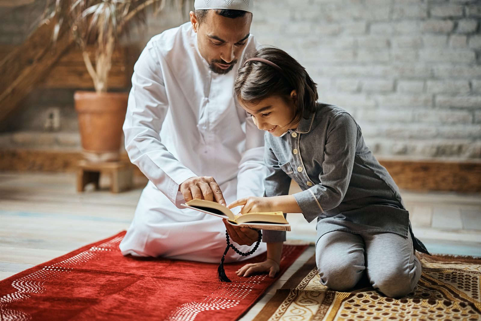 A father guiding his daughter through the Quran on a prayer mat