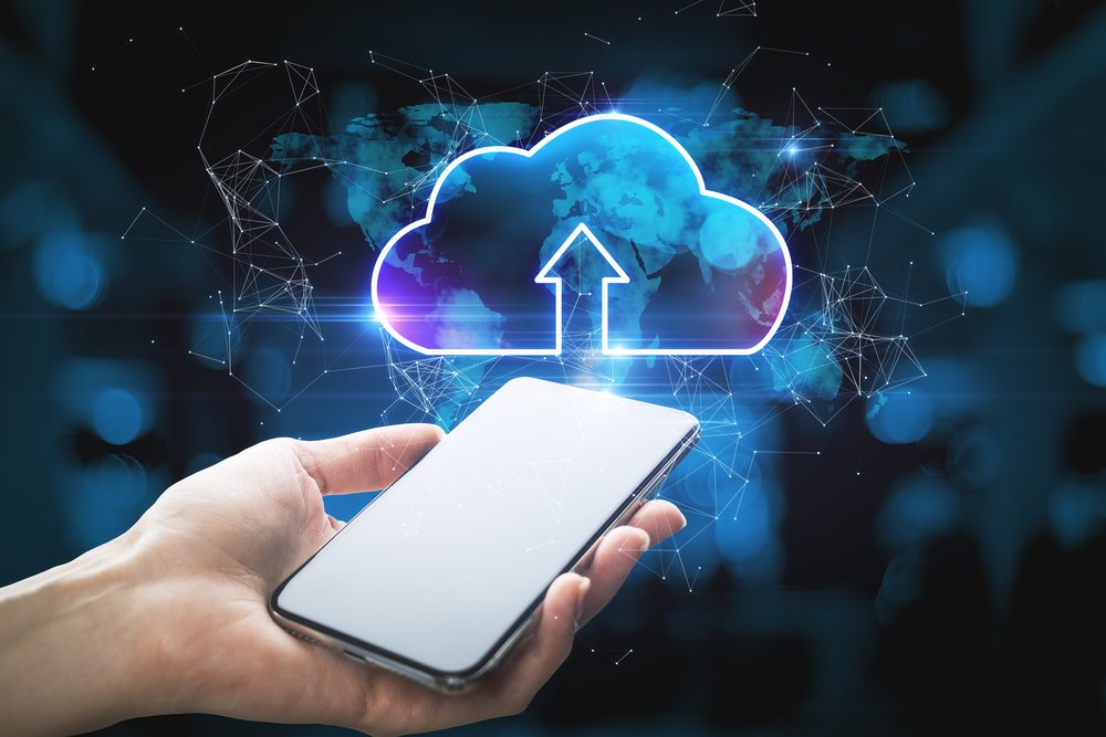 A hand holding a phone beneath a glowing cloud-upload icon over a world map